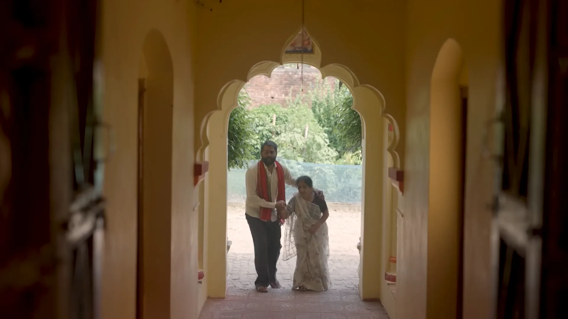 A son walks his mother through an arched doorway, from Jaise Maai Waise Betwa